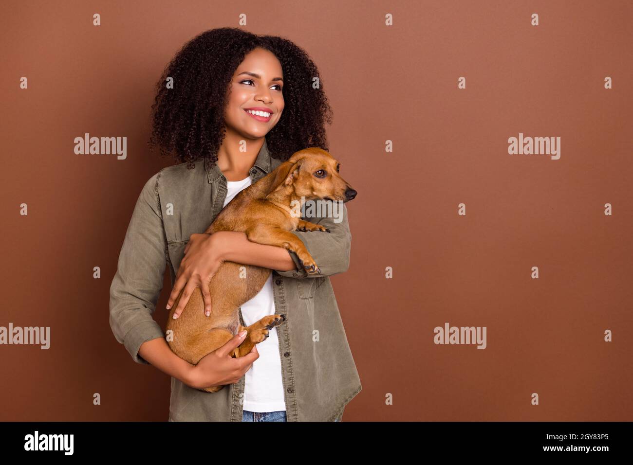 Photo of cheerful positive person arms hold pup look empty space toothy smile isolated on brown color background Stock Photo