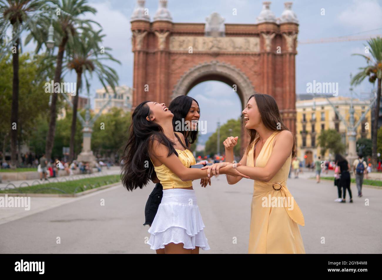 Stylish millennial multiracial girlfriends laughing brightly while standing together on urban street against triumphal arch during summer vacation in Barcelona city Stock Photo