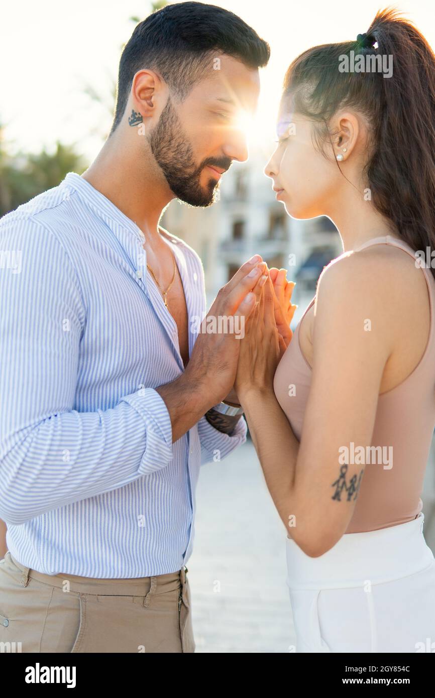 Side view of loving Hispanic couple gently holding hands while standing in city against bright sun Stock Photo