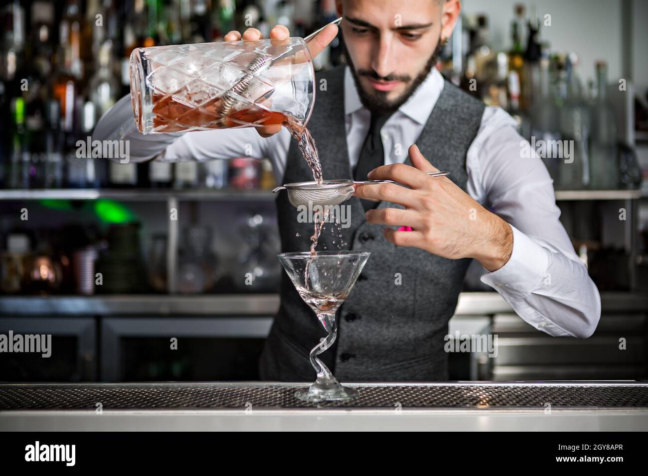 Elegant male barkeeper pouring alcohol drink through sieve into glass while preparing beverage at bar counter Stock Photo