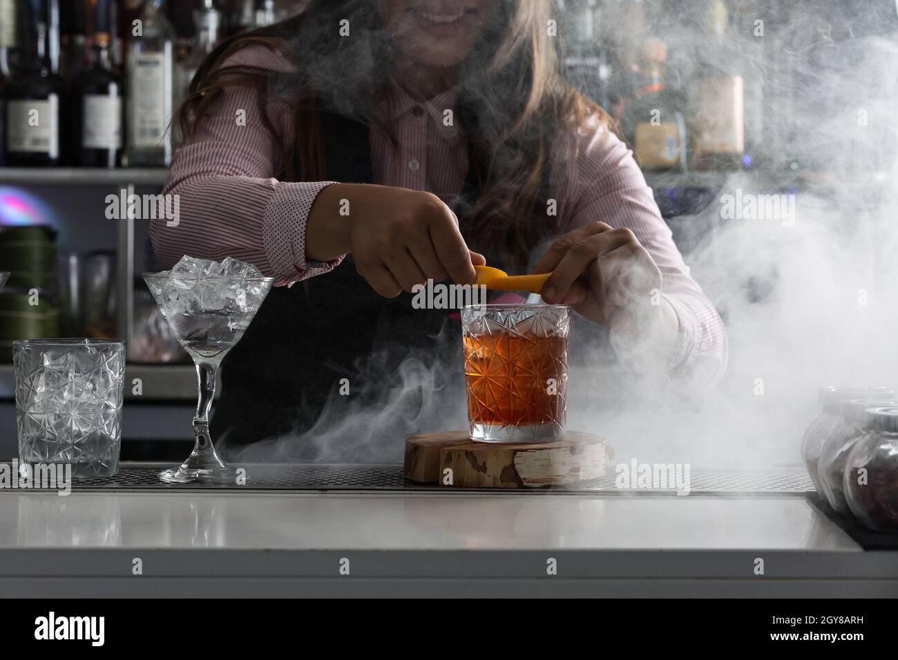 Crop cheerful female bartender adding fresh orange peel into glass with