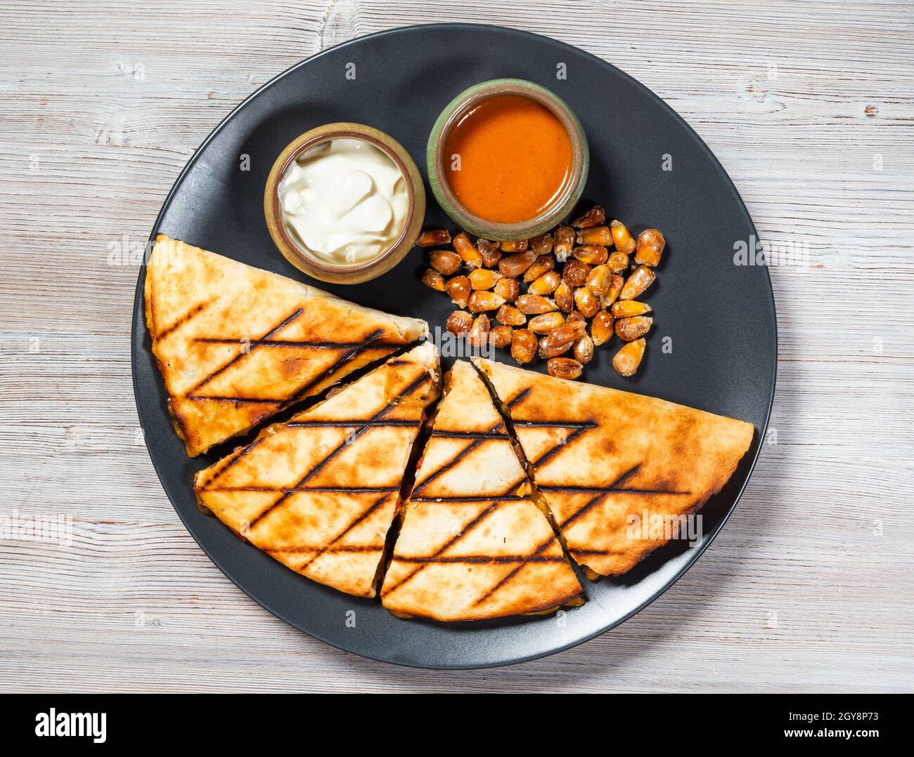 top view of cut Quesadilla from folded tortilla filled with cheese, meats, spices with sauces and fried corn seeds on plate on gray wooden board Stock Photo