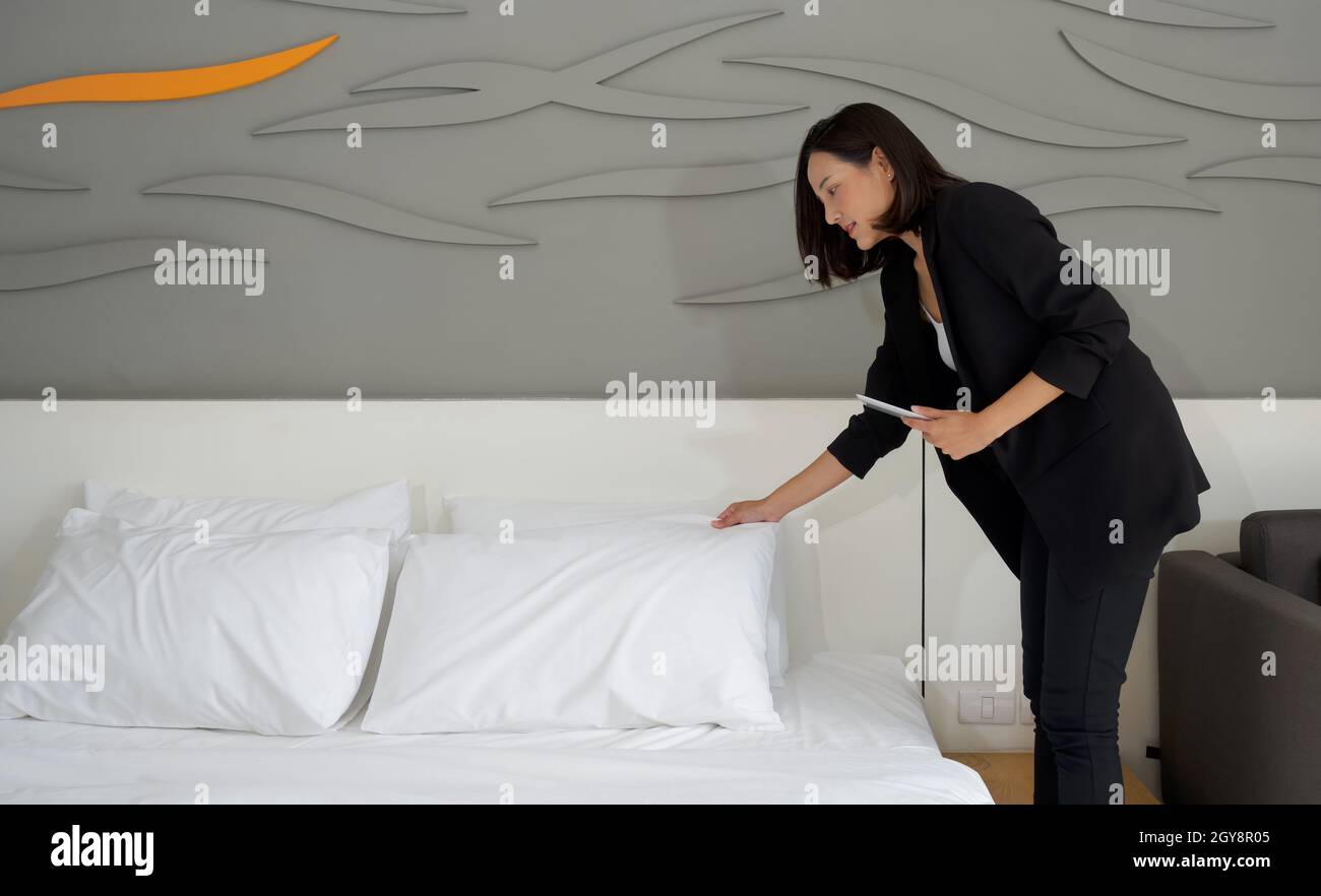 Asian businesswoman in black suit standing with tablet computer in her hand. The hotel manager verify the tidiness of the room for hotel guests. Stock Photo
