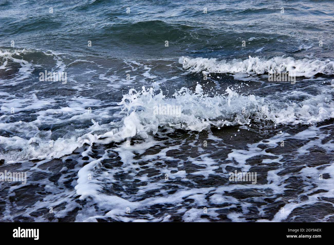 Splashes of sea foam. Dark sea, strong waves with bright white sea foam. The raging sea in bad weather. An incoming wave close up. Storm waves at sea. Stock Photo