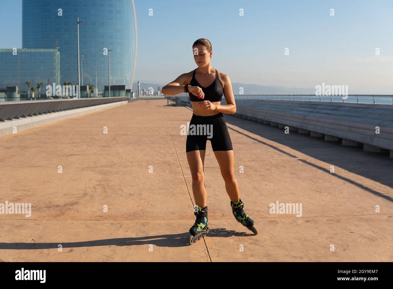 Full body female athlete in sportswear checking pulse on smart watch while riding roller skates on embankment in morning. Stock Photo
