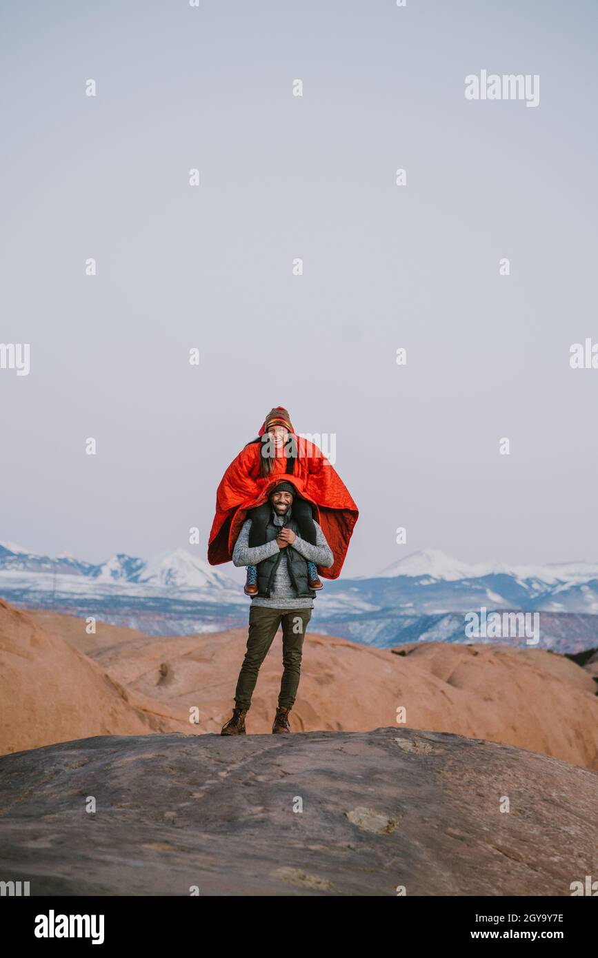 Young woman sits on top of her friend's shoulders wearing poncho in the mountains Stock Photo