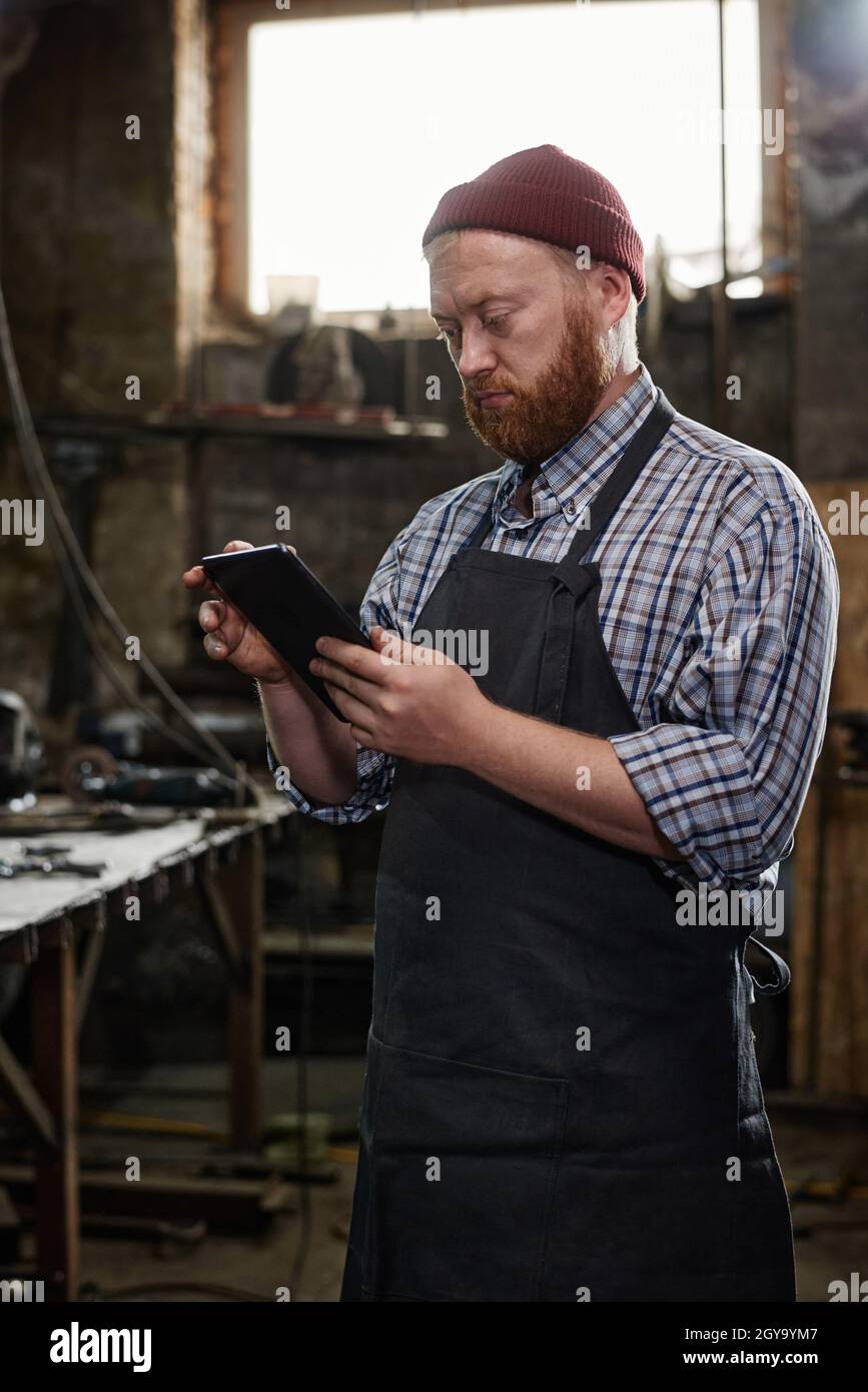Mature engineer in apron typing on digital tablet during his work in workshop Stock Photo