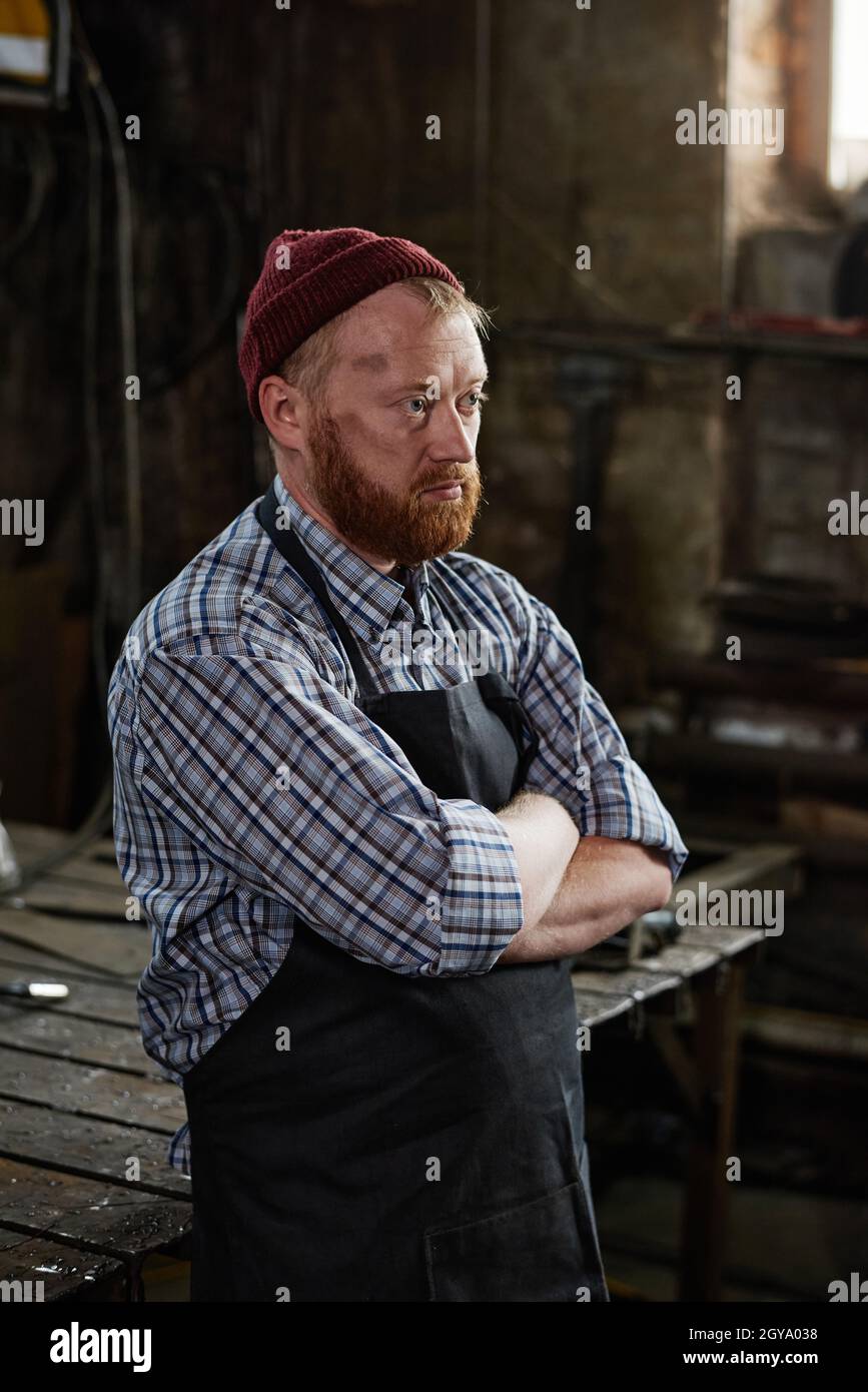 Mature man in apron standing with arms crossed and thinking about something in the workshop Stock Photo
