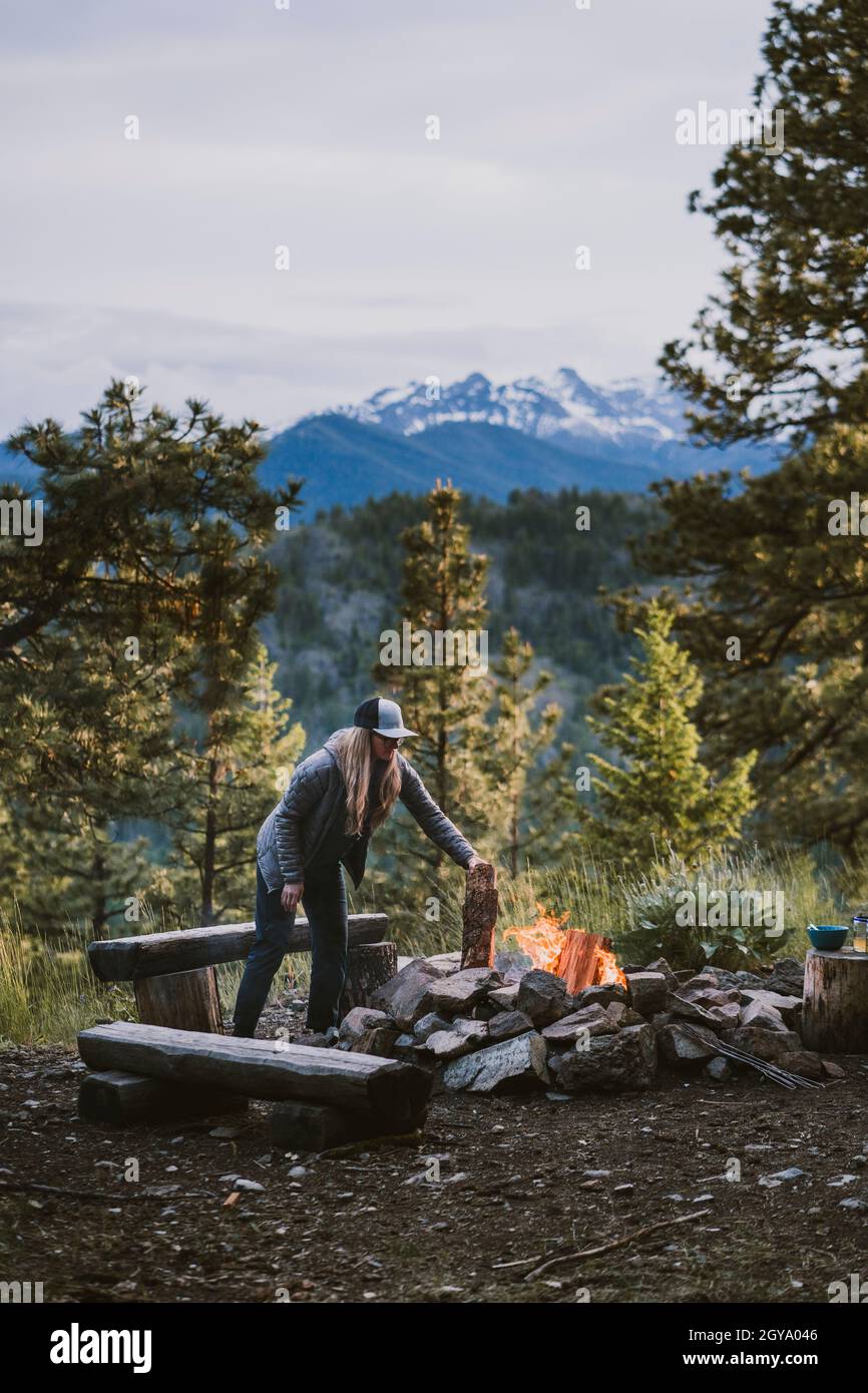 Woman wearing puffy jacket puts log onto campfire in the mountains Stock Photo