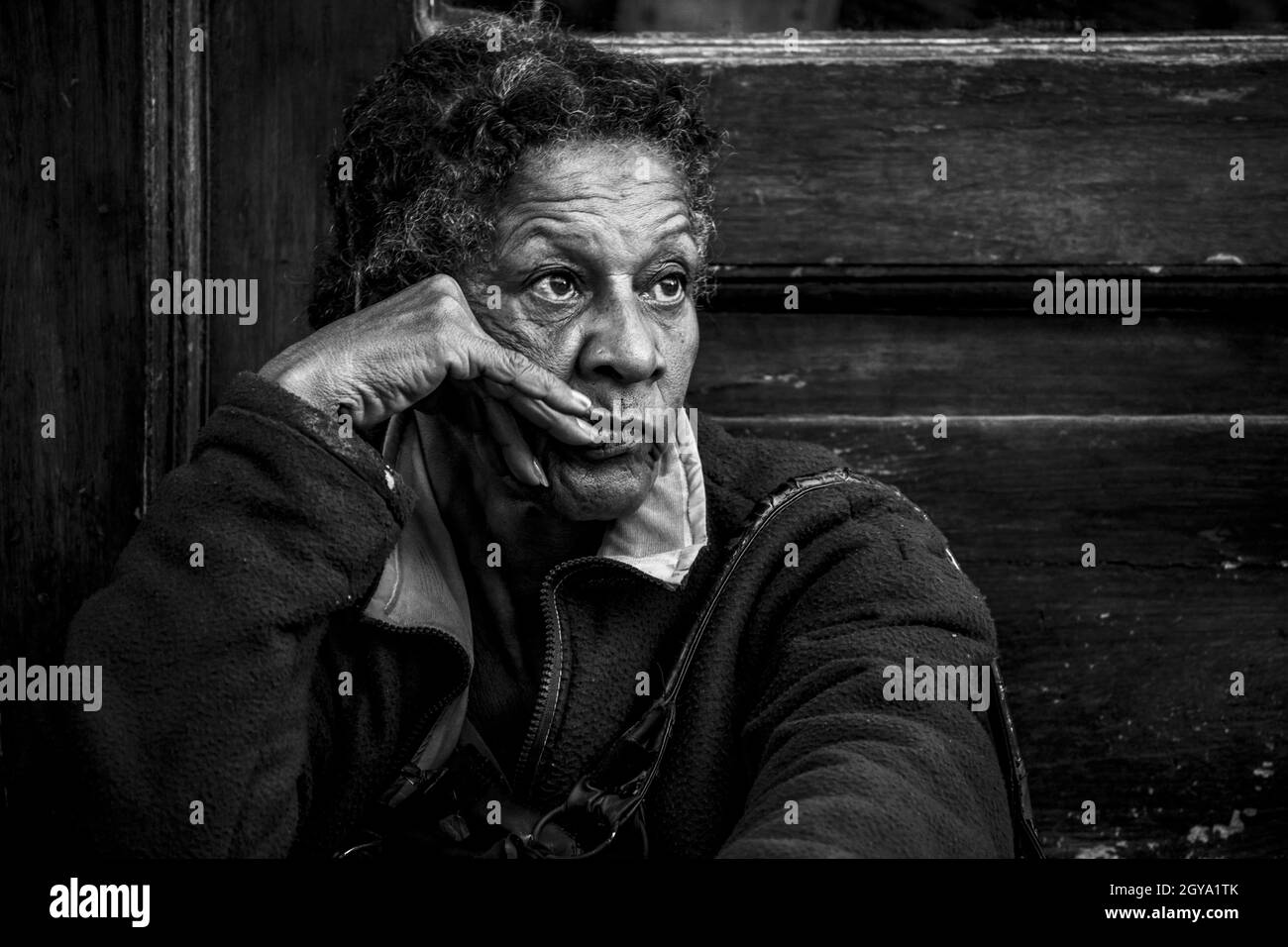 HABANA, CUBA - May 14, 2019: A grayscale shot of an old woman sitting in front of an old wooden background Stock Photo