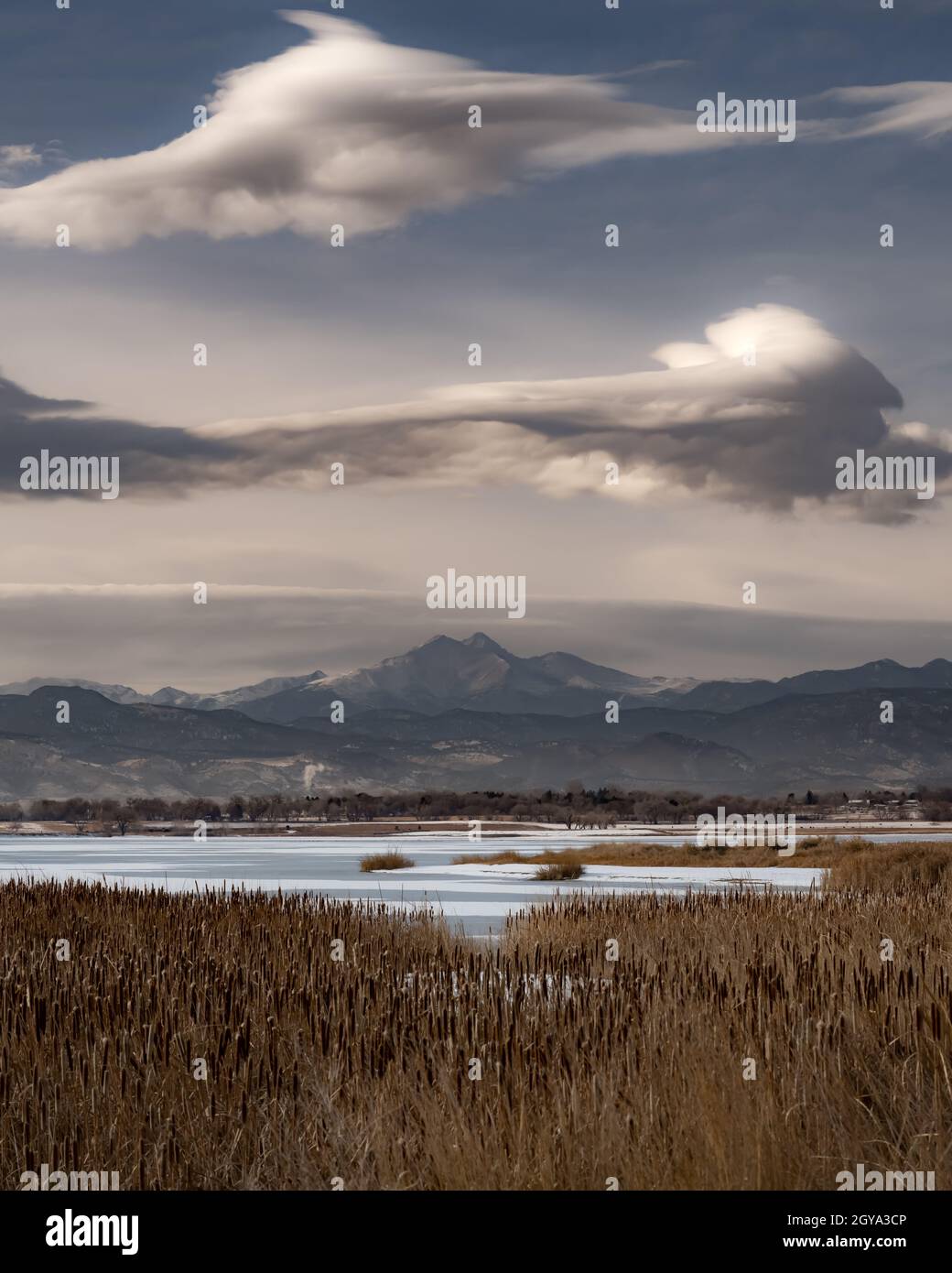 A mountain (Longs Peak) rises above a frozen lake on a cold winter's day. Unique clouds hang above the mountains, while reeds ling the lake. Stock Photo