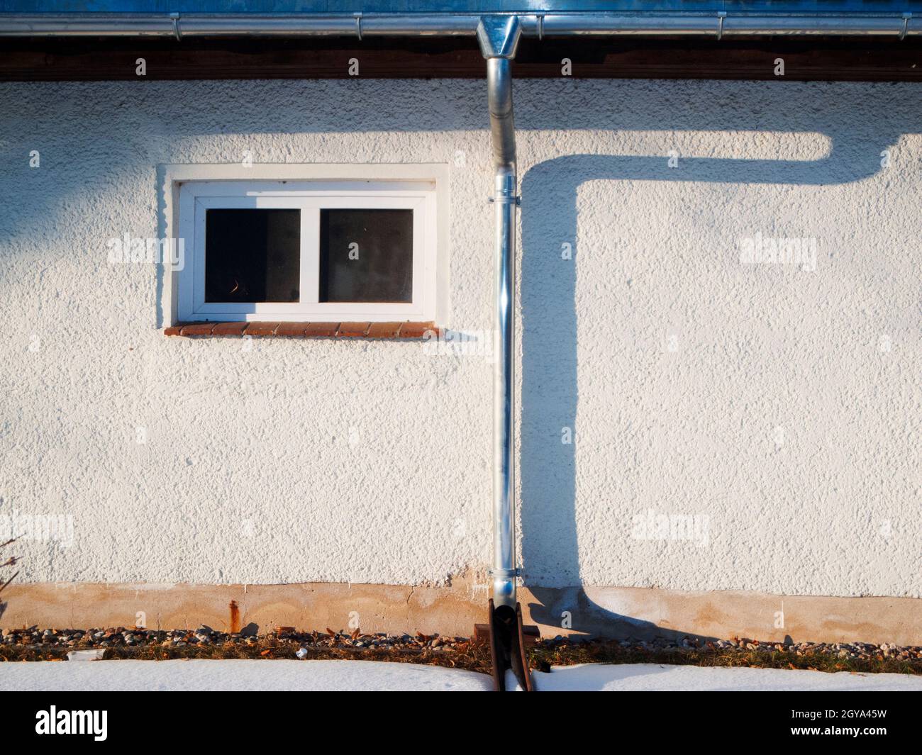 Detailansicht einer flachen Hausfassade mit Dachrinne, Fallrohr und kleines Fenster im seitlichen Abendlicht mit Schattenbild des Fallrohrs. Stock Photo