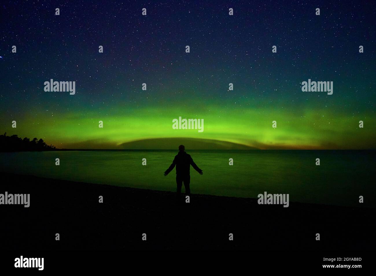 Man on edge of land before water lake ocean or sea with dome of green aurora light mist and tree silhouettes off to side Stock Photo