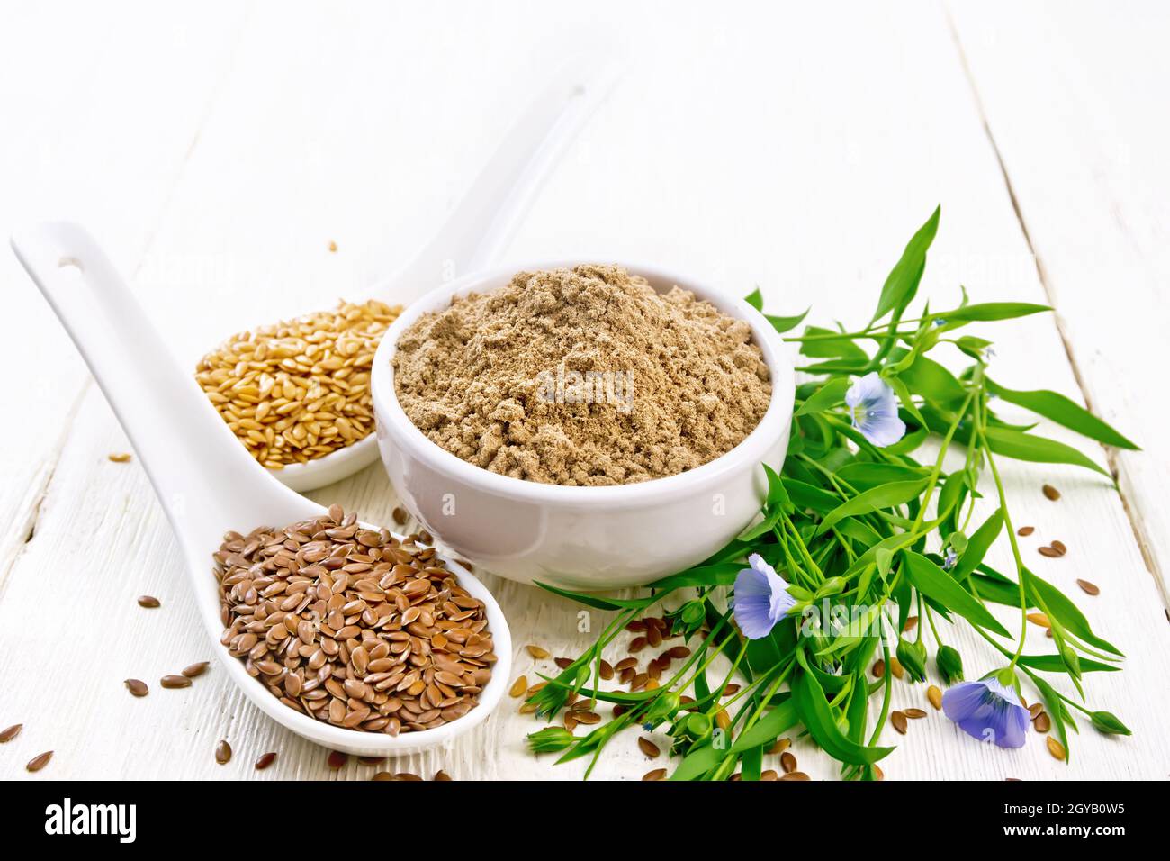 Flaxseed flour in a bowl, white and brown linen seeds in two spoons and on table, flax leaves and flowers on white wooden board background Stock Photo