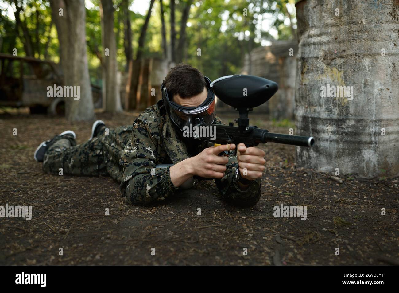 Male paintball player with gun shoots lying down on the ground