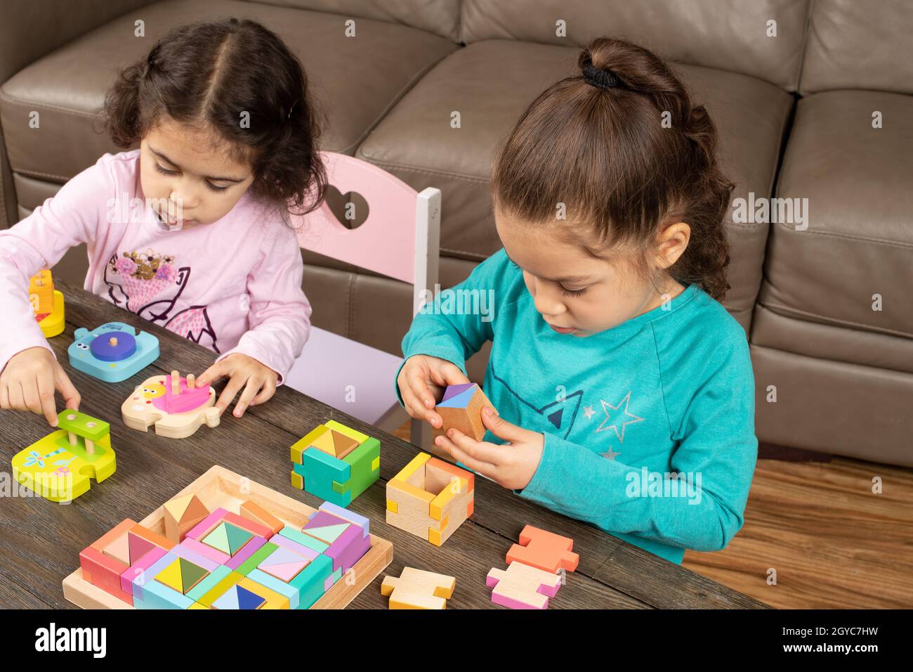 Five year old girl working on complex geometric shapes activity while three year old sister plays next to her with simpler geometric shapes puzzle Stock Photo