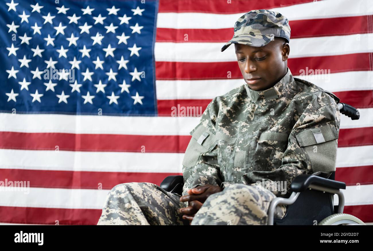 Patriotic Soldier Sitting In Wheel Chair Against American Flag Stock Photo