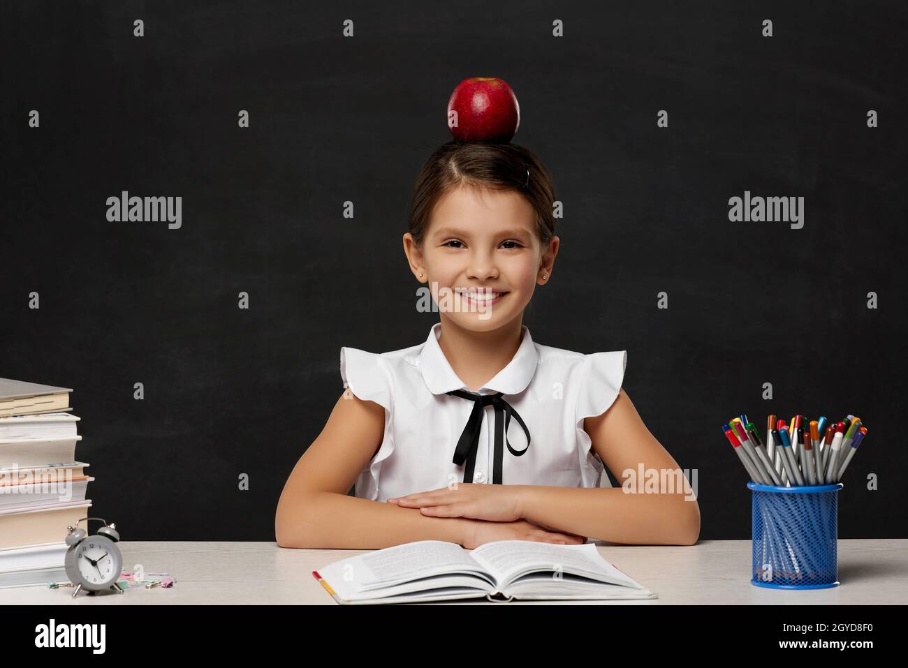 little child girl studying in classroom on background of blackboard. schoolgirl with red apple on her head. Back to school. Stock Photo