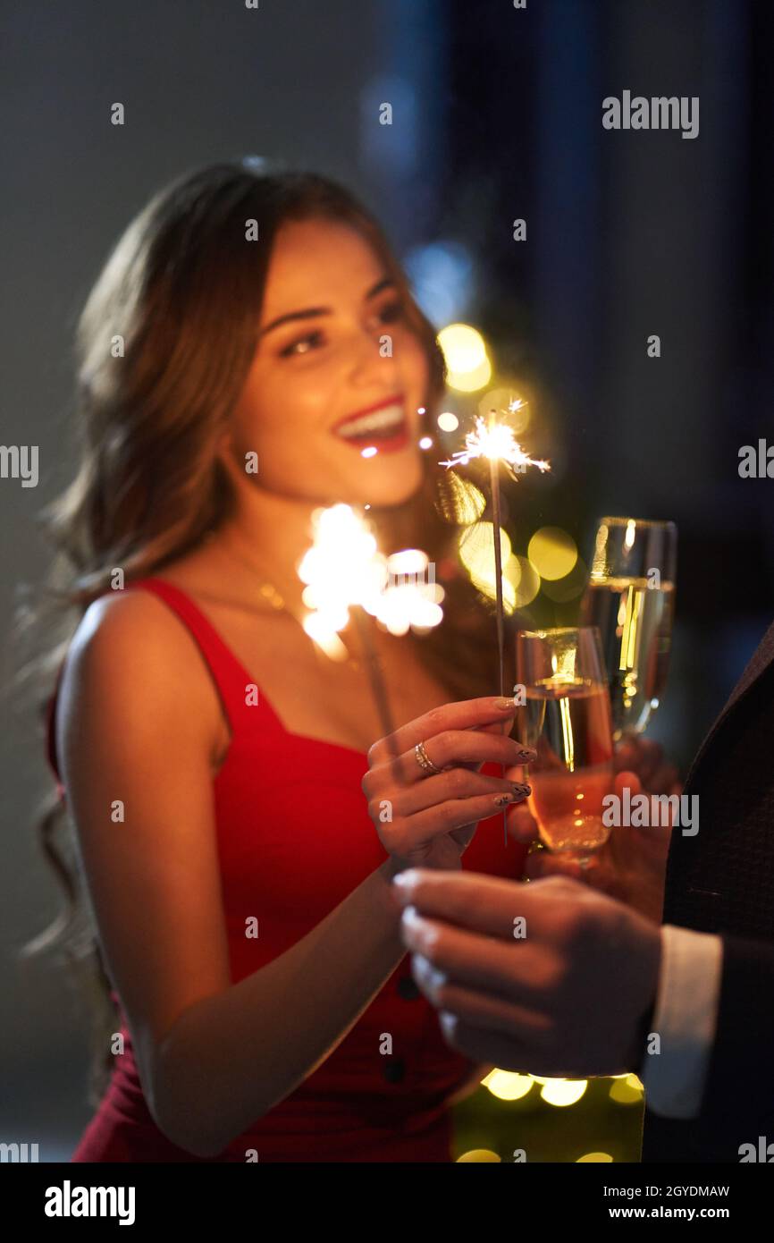 Attractive caucasian woman in red dress celebrating new year with her lovely man. Happy couple drinking champagne and holding sparklers. Holiday mood. Stock Photo