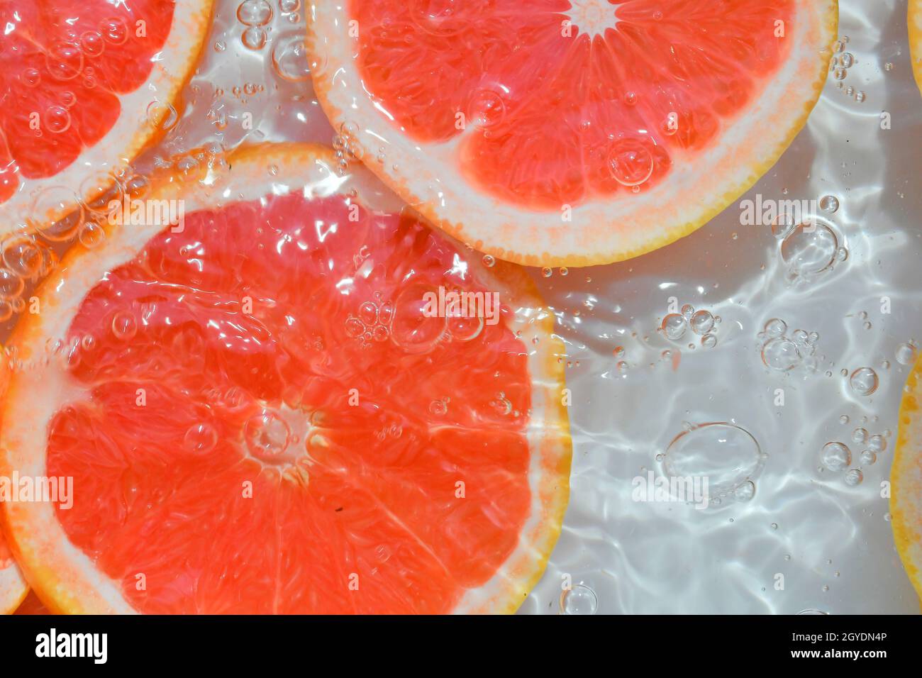 Grapefruit close-up in liquid with bubbles. Slice of blood red ripe grapefruit in water. Close-up fresh slices of red grapefruit on white background. Stock Photo