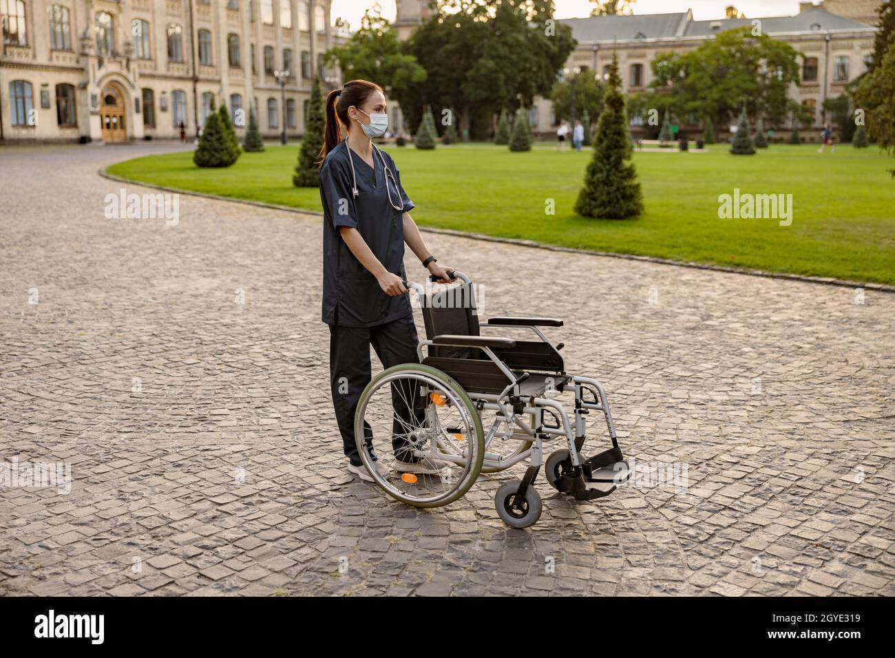 Full length shot of caring young nurse wearing protective mask during Covid19 pandemic standing with wheelchair outdoors in the park near clinic Stock Photo
