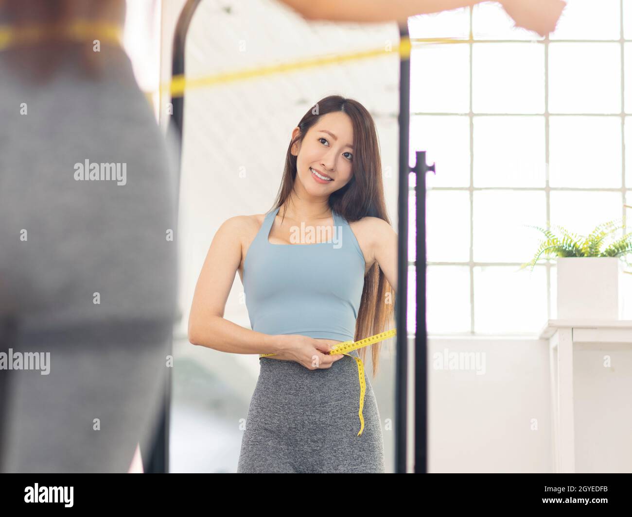 Young woman measuring waist with tape standing in front of mirror