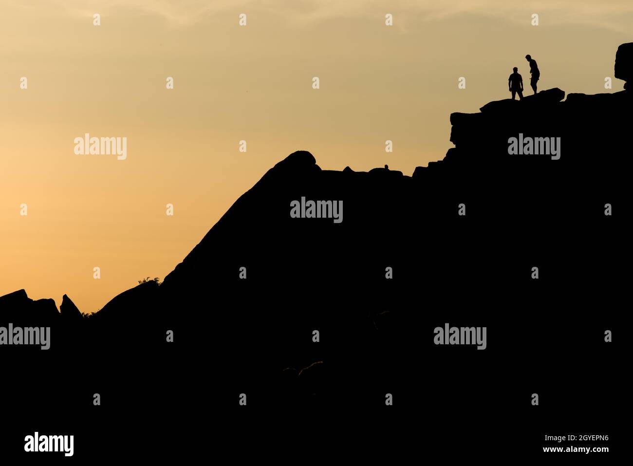 A pair of rock climbers, silhouetted at sunset, look back from the summit of stange edge. Located in the peak district in england. Outdoor activity Stock Photo
