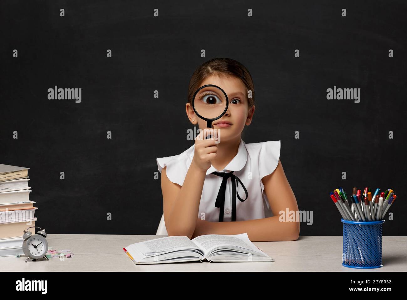 cute happy schoolgirl looking through magnifying glass in the classroom on blackboard background