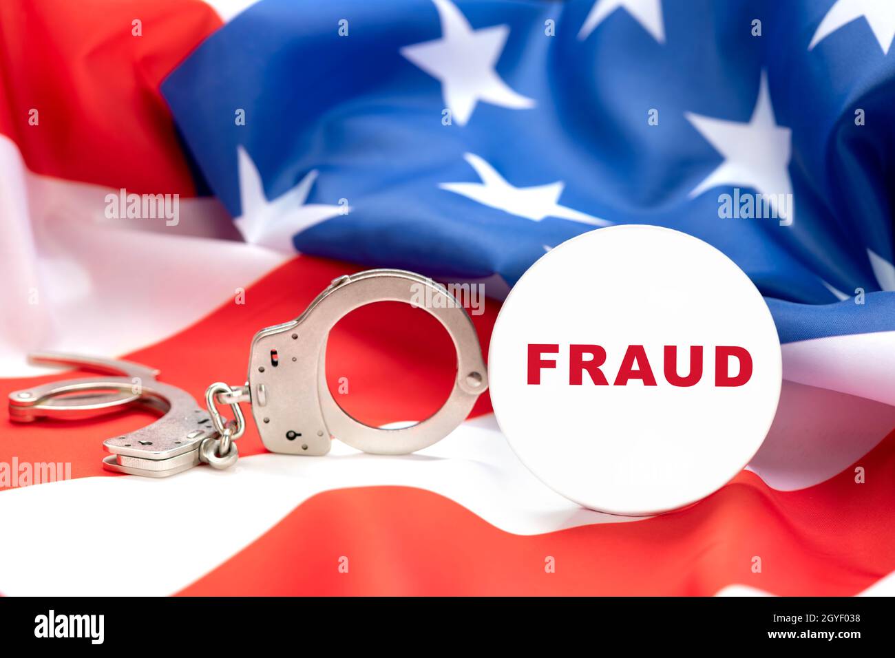 Images depicts a fraud button against handcuffs and the American flag, insinuating that fraud is a crime and those cheating people will be arrested. Stock Photo