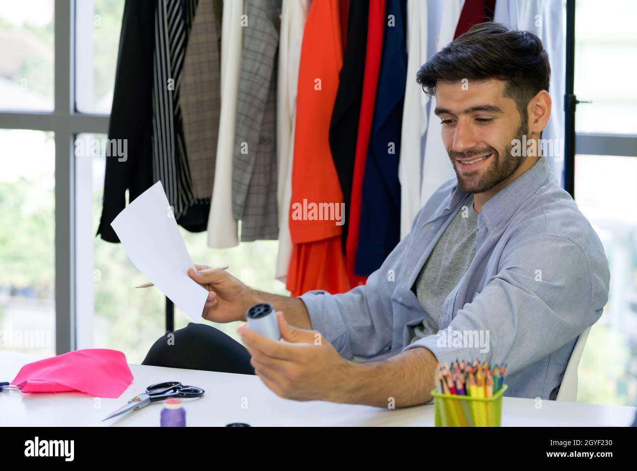 The dressmaker is satisfied with the color of the yarn used to design the new shirt for the customer. A clothes rack full of suits is placed in front Stock Photo