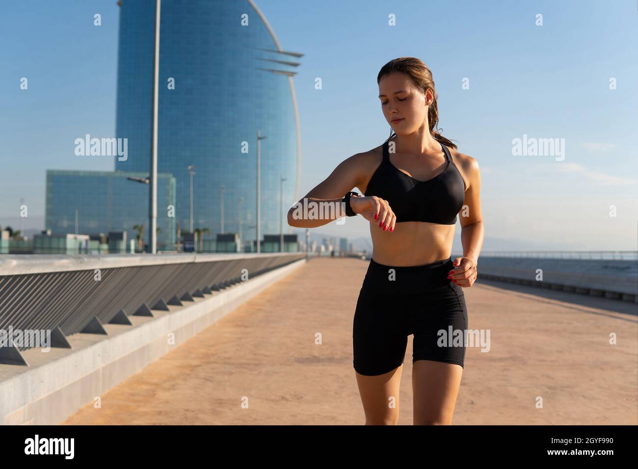 Fit sportswoman in sports bra and shorts running and checking pulse on smart band during cardio workout on embankment in city. Stock Photo