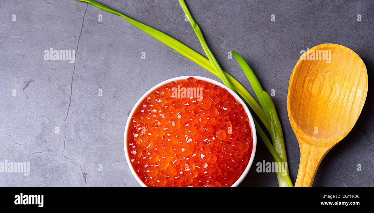 Red caviar in a wooden cup on a gray background with a spoon and green onions. A large pile of bright caviar. Fresh delicious caviar. A place for adve Stock Photo