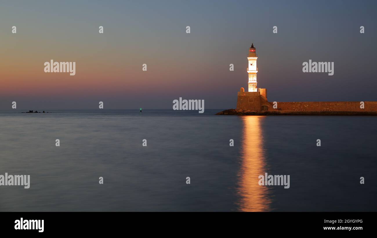 Panoramic view with lit lighthouse in Chania, Crete, Greece, reflections in water, horizon, beautiful early morning sunrise light Stock Photo