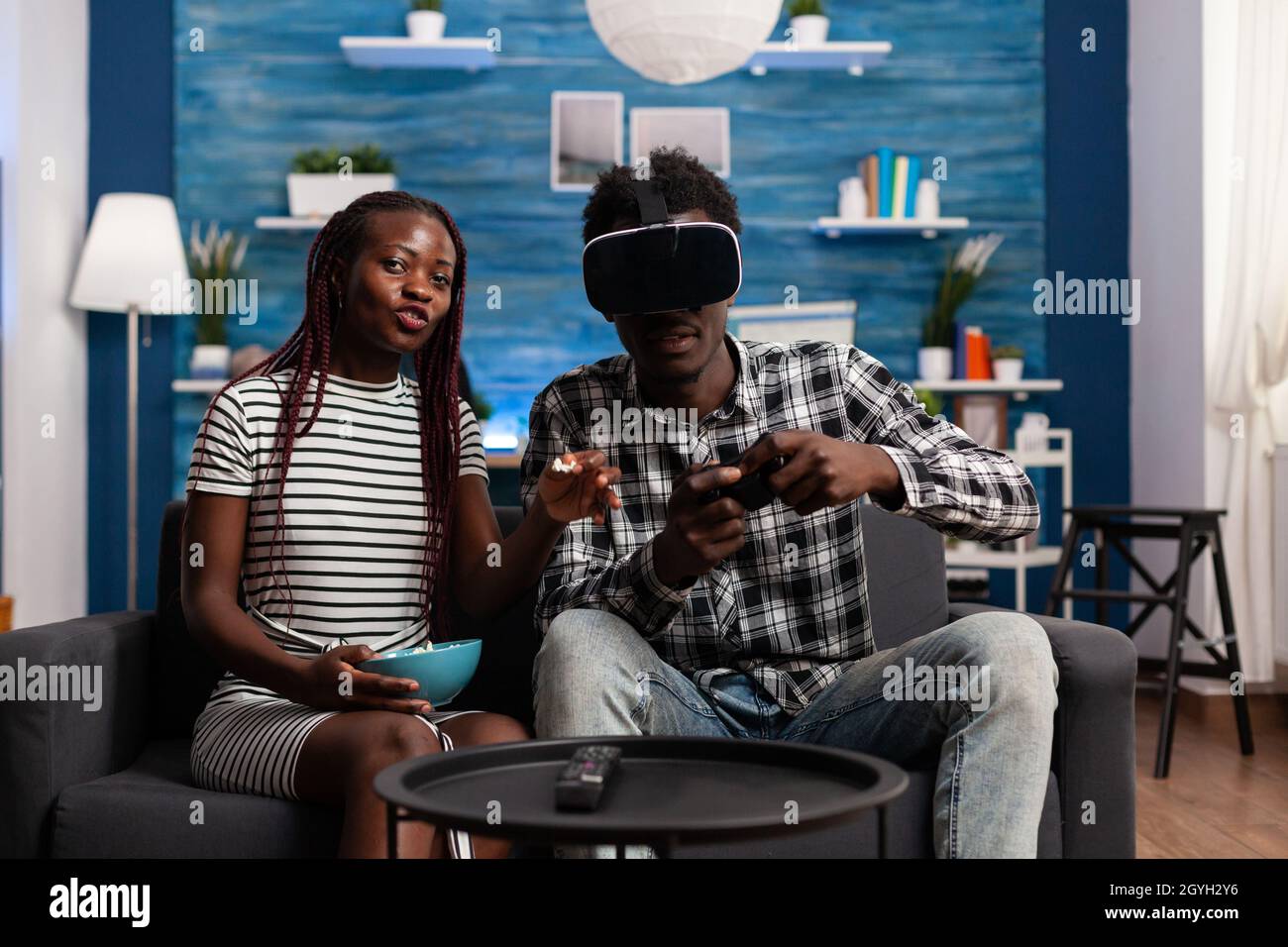 POV of black couple using technology for entertainment at home. African  american man wearing vr glasses headset and playing with joystick while  young woman watching video game on television Stock Photo -