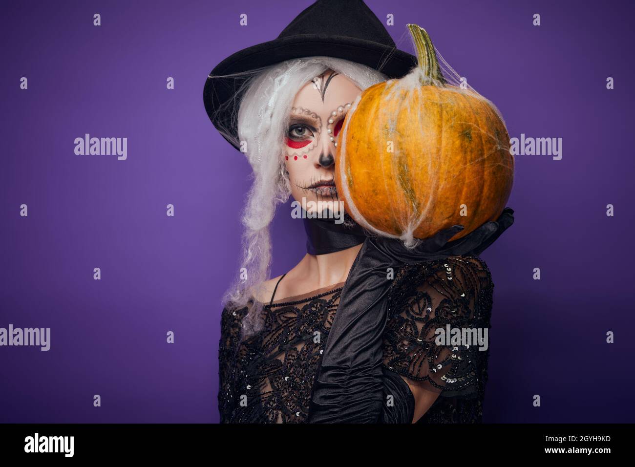 Serious young woman with Halloween make-up covers half of face with pumpkin Stock Photo