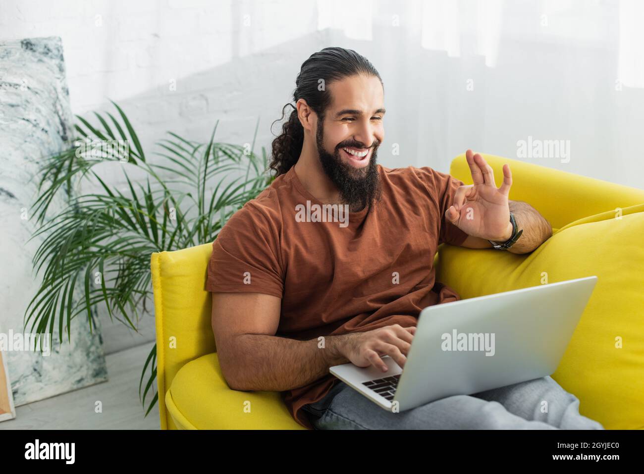 cheerful latin man showing okay gesture while using laptop on sofa at ...
