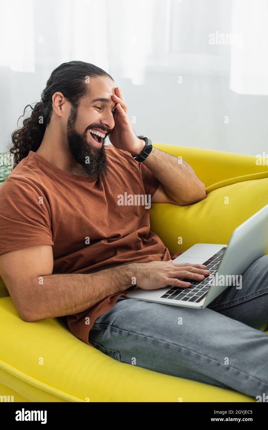 excited hispanic man laughing while using laptop on couch at home Stock Photo