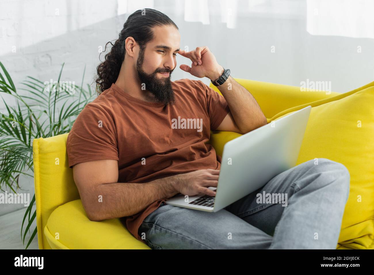 positive and thoughtful hispanic man looking at laptop while sitting on sofa at home Stock Photo