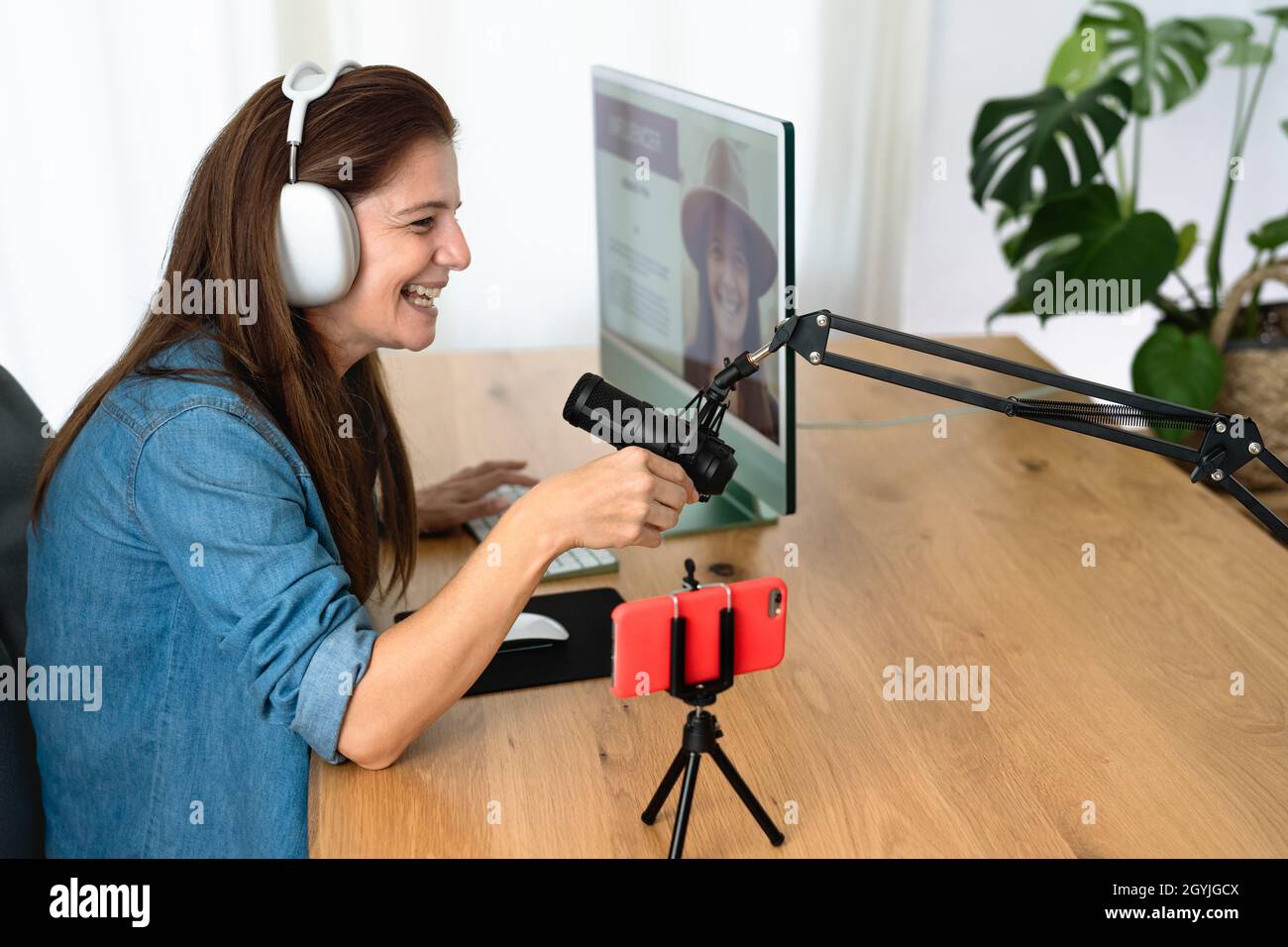 Mature woman recording a podcast using microphone and laptop while streaming on social network with mobile smartphone from her home studio Stock Photo