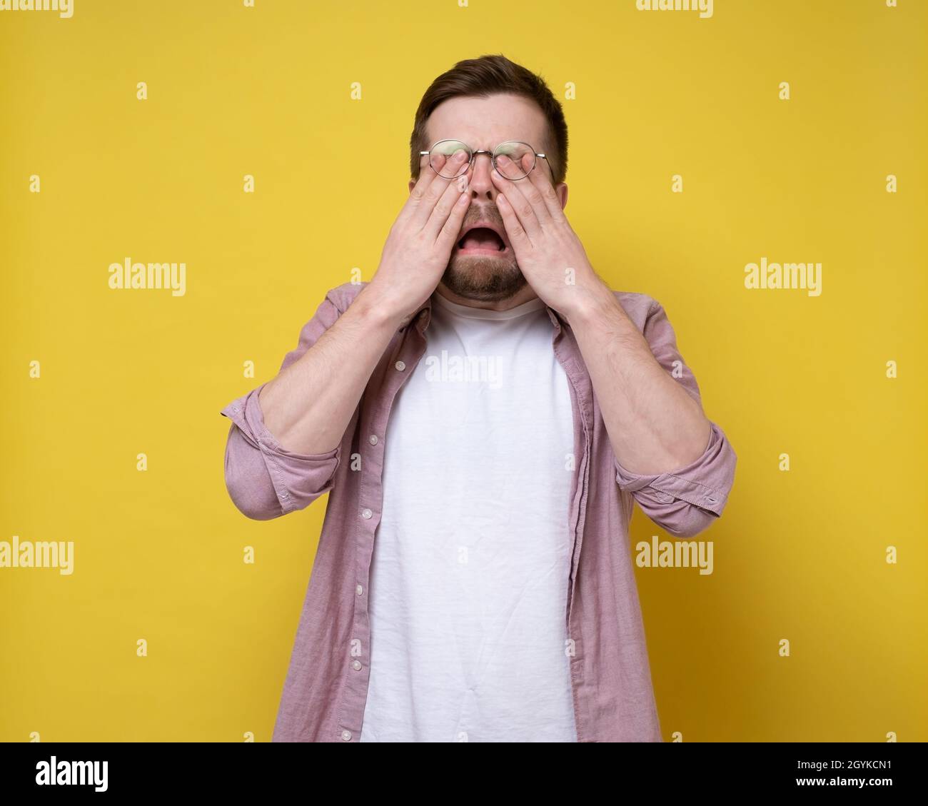 Unhappy Caucasian man is upset and crying, he wipes eyes with hands and opens mouth in sobs. Yellow background. Stock Photo
