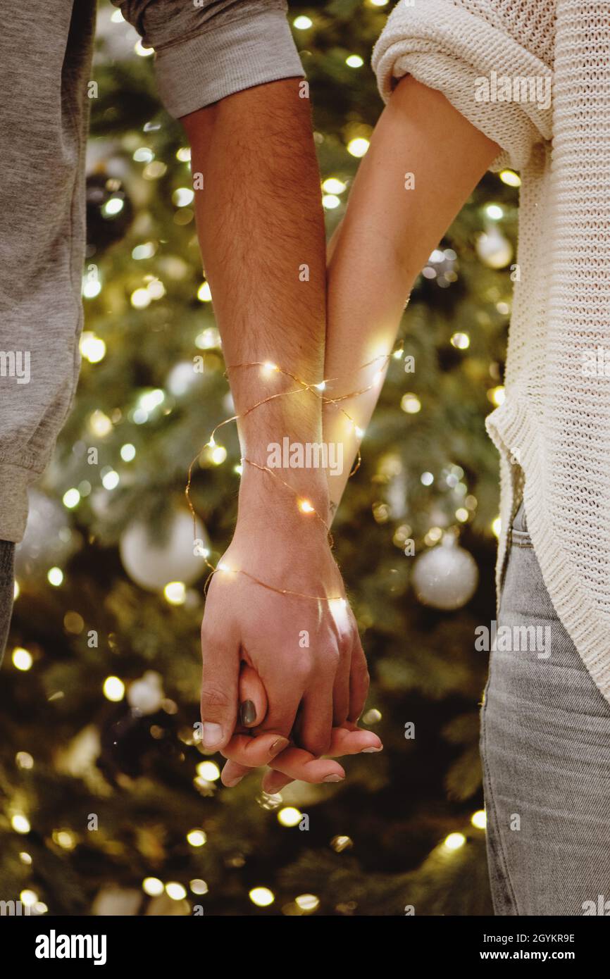 Partial view of couple holding hands wrapped in a garland with Christmas tree on background. Stock Photo
