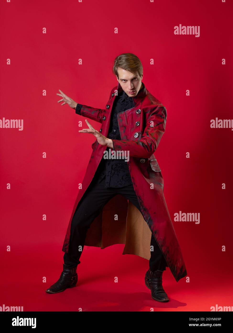 An outrageous young man in a daring red coat in a vintage noir style, epic poses and makes a hand gesture. photo on a red background Stock Photo