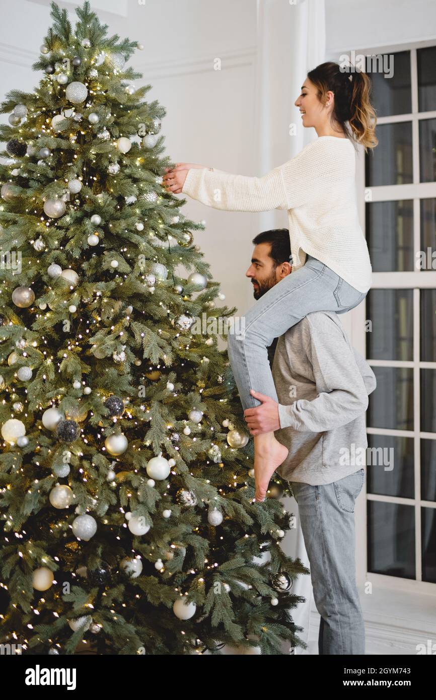 Couple decorating Christmas tree. Woman sitting on the shoulders of man. Stock Photo