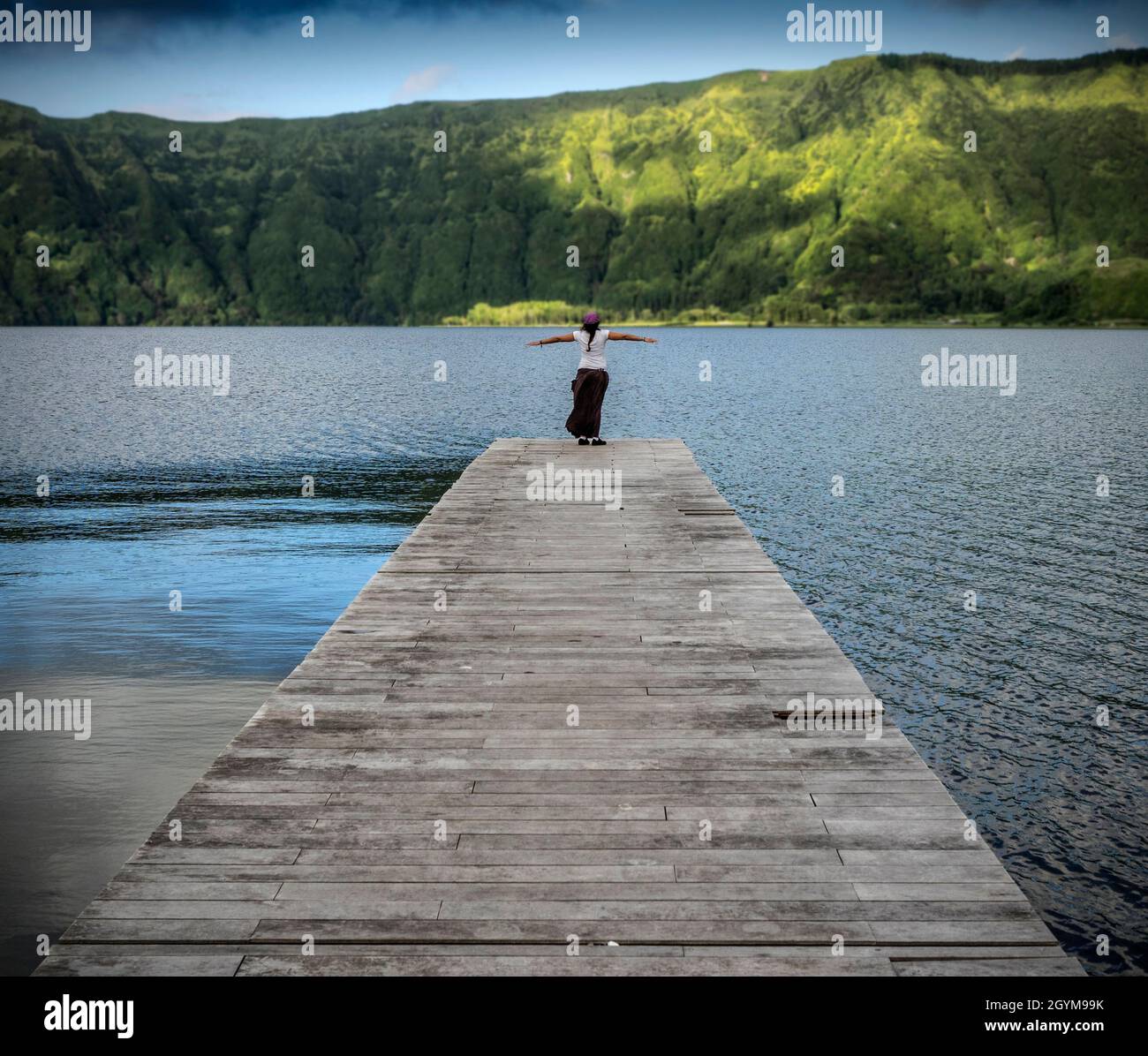 A young girl with open arms at the end of a wooden jetty in a lake in the Azores. Stock Photo