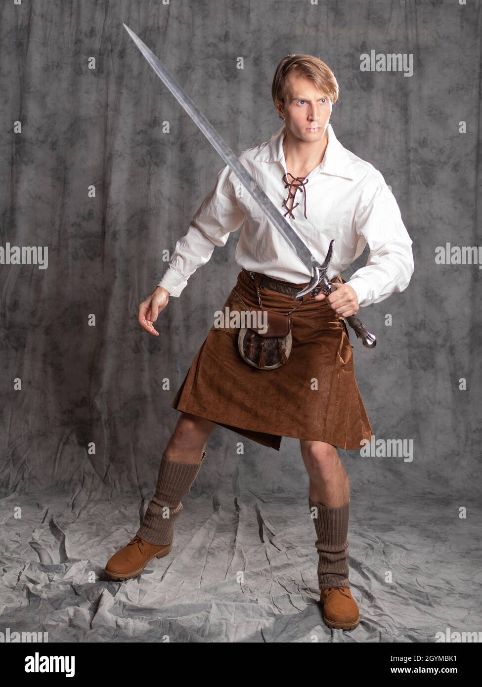 A young man in a leather kilt and a white lace-up blouse. combat stance. A Scottish knight with a two-handed sword. Photo in the studio on a gray back Stock Photo
