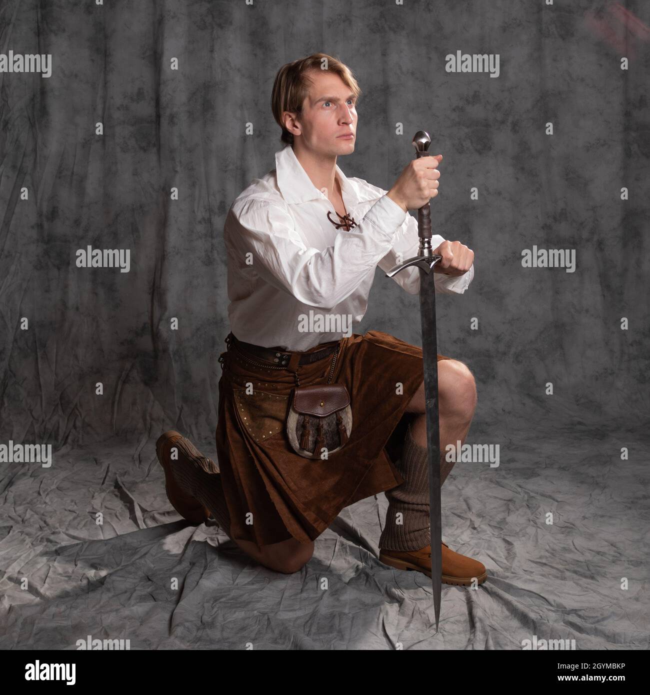 The Scottish knight knelt, took an oath, concept. A young man in a kilt with a sword in his hands in a pose on one knee, photo in the studio on a gray Stock Photo
