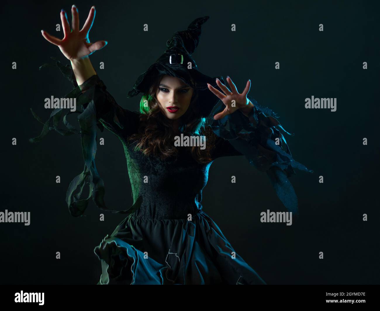 A charming young witch in a costume makes magical passes with two hands, a caster. Young brunette in pointed hat and black dress, photo on black Stock Photo