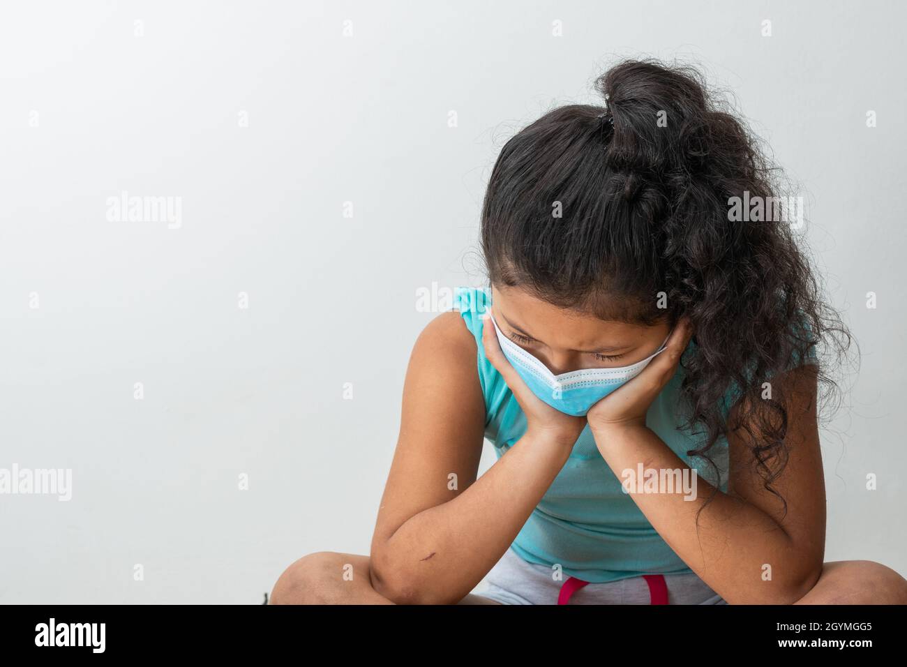 little girl (8 years old) sitting on the floor in anger, her hands are over her face and she has a blue surgical mask. Medical, pharmaceutical and san Stock Photo