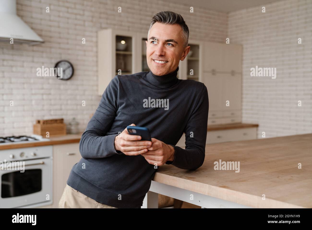 Handsome smiling man using mobile phone while leaning on table at home kitchen Stock Photo