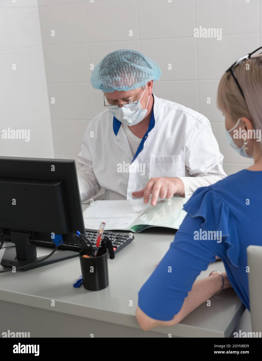 A doctor in a white coat, mask and cap in the office at the table reads the patients medical history Stock Photo